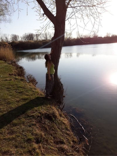 Frau im 5. Monat schwanger lehnt an Baum am See mit Sonnenschein